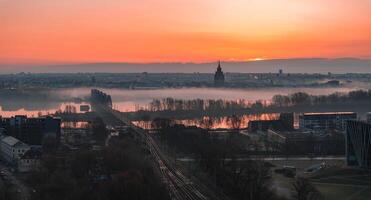 Serene sunrise over Riga, Latvia, featuring St. Peter's Church spire, mist covered Daugava River, a railway bridge, and urban structures in soft light. photo