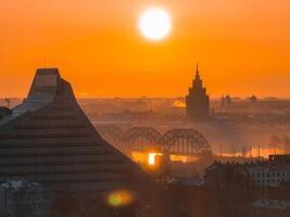 The image shows a sunrise over Riga, Latvia, featuring the National Library, the Latvian Academy of Sciences, the Daugava River, and a railway bridge. photo