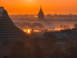 A warm sunrise illuminates Riga, Latvia, featuring the arched railway bridge over the Daugava River, the Latvian Academy of Sciences, and the National Library. photo