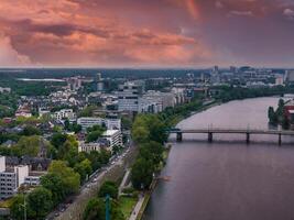 Aerial image of Frankfurt, Germany, featuring the Main River, a bridge, urban skyline, lush greenery, and a vibrant sunset with a faint rainbow. photo