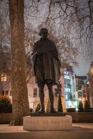 A statue of Mahatma Gandhi on a pedestal surrounded by bare trees, illuminated buildings, and streetlights in a London outdoor setting at night. photo