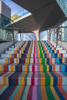 Vibrant staircase with bold striped patterns, glass railings, and a modern structure. A tree and building reflections add depth to the urban scene. photo