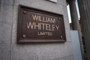 A bronze plaque engraved with William Whiteley Limited is mounted on a light gray stone wall, with part of a column and a building in the background. photo