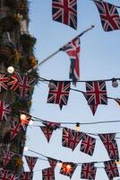 Union Jack flags and glowing string lights hang against a clear blue sky. A building with colorful flower arrangements adds greenery and vibrancy to the scene. photo