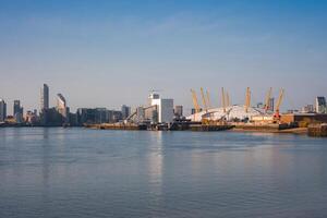 The O2 Arena's white dome and yellow masts stand out along the River Thames, surrounded by modern buildings. The Monier Wharf sign is visible under a clear sky. photo