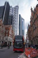 A red double decker bus with Shoreditch 78 displayed travels on a London street, surrounded by modern skyscrapers, red brick buildings, and pedestrians. photo