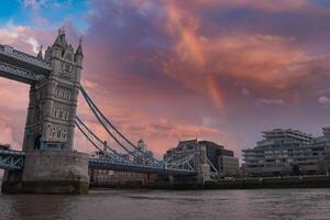 Tower Bridge in London stands against a vibrant sunset with orange and pink hues. A rainbow arches above, and the Thames River flows in the foreground. photo