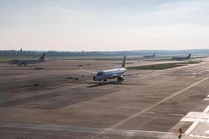 An airport runway with multiple airplanes, including one taxiing in the foreground. A spire or tower is faintly visible on the distant horizon. photo
