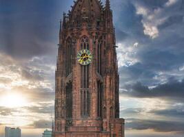 The Gothic style tower of Frankfurt Cathedral with intricate red sandstone details, a clock face, and a glowing sunset sky. Modern buildings in the distance. photo