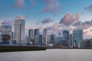 Modern skyscrapers of Canary Wharf in London with the River Thames in the foreground, construction cranes, and a partly cloudy early evening sky. photo