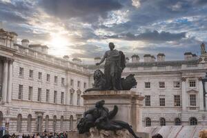 A statue with a lion and reclining figures stands in Somerset House courtyard, London, with neoclassical architecture and a dramatic sky in the background. photo