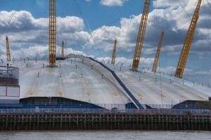 The O2 Arena in London, England, features its white dome and yellow masts. The River Thames flows in the foreground under a partly cloudy sky. photo