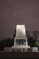 The Guards Memorial in London stands illuminated at night, with bronze soldier statues, detailed inscriptions, wreaths, and trees in the background. photo