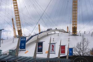 The O2 Arena in London features climbers on its white dome with yellow masts, brand banners in the foreground, and an overcast sky in the background. photo