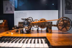 A historical machine with intricate gears, wheels, and levers on a wooden surface. A keyboard in the foreground suggests a musical connection. Museum setting. photo