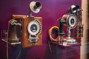 A wooden rotary telephone with a crank handle and a polished metallic telephone with ornate detailing are showcased in a curated collection. photo