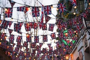 A London street decorated with Union Jack flags and warm string lights. Traditional architecture, bare trees, and greenery enhance the urban scene. photo
