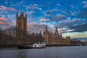 The Palace of Westminster with Gothic architecture stands by the River Thames at sunset. Boats are moored in the foreground under a partly cloudy sky. photo