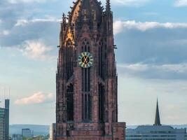 The Gothic style tower of Frankfurt Cathedral with intricate details, a gold accented clock, and modern buildings in the background under a cloudy sky. photo
