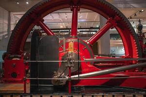 A red industrial steam engine with a massive flywheel and connecting rods, displayed indoors with protective railings in a modern London museum. photo