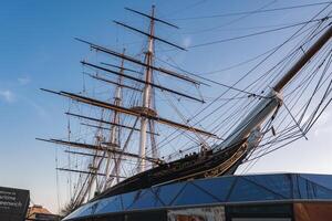 The Cutty Sark, a 19th century clipper ship in Greenwich, London, displays tall masts and rigging above a modern glass structure under a clear blue sky. photo