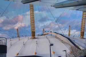 The O2 Arena in London features its white dome, yellow masts, and visible walkway. Modern buildings frame the scene under a blue and pink sky. photo