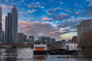 Serene view of the River Thames in London with moored boats, a glass skyscraper, a curved residential building, and a bridge under a colorful sky. photo