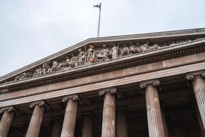 The facade of a neoclassical building in London features a triangular pediment with sculptures, fluted columns, and a Union Jack flag under a cloudy sky. photo