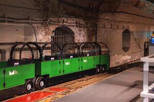 A green miniature train with transparent domed roofs sits on tracks in a brick lined underground tunnel, part of the historic Mail Rail in London. photo
