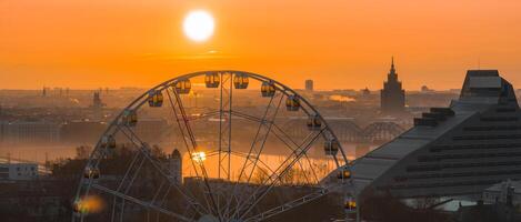 A sunrise view of Riga, Latvia, featuring a Ferris wheel, the National Library, the Latvian Academy of Sciences spire, and bridges over the Daugava River. photo