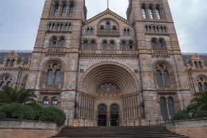 The grand facade of the Natural History Museum in London features Romanesque architecture, arched windows, tall towers, and greenery in the foreground. photo
