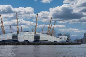 The O2 Arena's white dome and yellow masts stand by the River Thames, with Canary Wharf skyscrapers in the background under a partly cloudy sky. photo