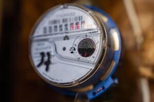 Close up view of a water meter with a clear casing, showing a white dial, black and red counters, a red gauge, and visible brand and model details. photo