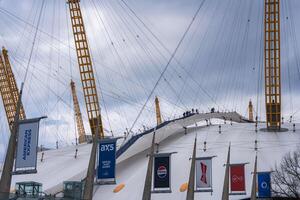 The O2 Arena in London features its iconic white dome, yellow masts, climbers on top, branded banners in the foreground, and a cloudy sky backdrop. photo