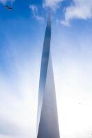 A sleek metallic structure with a pointed design rises against a bright blue sky with scattered clouds. A bird is in flight near the top left corner. photo
