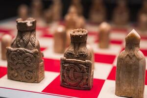 Chess pieces with ornate medieval style designs, including a crowned king and bishop, displayed on a red and white board in an exhibition setting. photo
