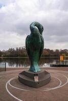 A green bronze bird sculpture on a circular pedestal near a calm lake with a swan, dock, autumn trees, and an overcast sky in a London park. photo