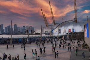 The O2 Arena's white dome and yellow towers stand out in London, with a busy plaza in the foreground and Canary Wharf's skyline in the background. photo