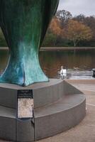 A green patinated sculpture on a stone pedestal with a plaque near a calm body of water, a white swan, and autumn colored trees in a London park. photo