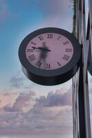 A round pink clock with Roman numerals is mounted on a reflective modern building. Shadows from the clock hands and soft clouds enhance the scene. photo