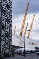 The O2 Arena's white dome and yellow masts stand near a modern building with circular windows, a paved pedestrian area, trees, and a cloudy sky. photo