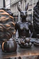 A bronze sculpture of a rabbit, dog, and lion around a table with a teapot and fruit, set against historic style buildings and black iron fencing in London. photo