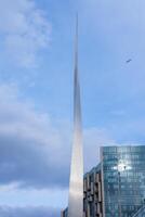A sleek metallic spire rises against a partly cloudy blue sky, with a modern glass building in the foreground and a bird in flight overhead. photo