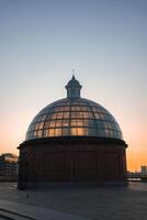 The domed glass and brick entrance of the Greenwich Foot Tunnel is lit by sunset hues. Modern buildings and the River Thames are visible in the background. photo