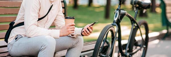 Work outdoors, take a break and get to office in morning. Smiling millennial man manager sits on bench with bicycle, holds takeaway paper cup of coffee and reads message in gadget, in city, outdoor photo