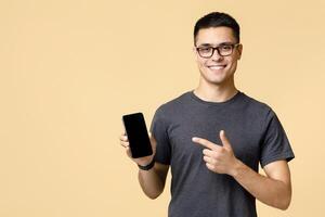 Blogger insists at better offer for customers. Happy handsome young guy in brown t-shirt and glasses, points finger at mobile phone with blank screen advertising app, isolated on beige background photo