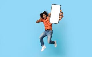 Excited young african american guy in casual jumping up with smartphone and pointing at white empty screen, recommending nice mobile app, blue studio background, mockup, copy space, full length photo