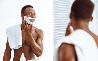 Shirtless African Guy Applying Shave Foam On Face Preparing For Shaving Standing Near Mirror In Bathroom Indoors. Male Facial Skincare And Selfcare Routine Concept. Selective Focus photo