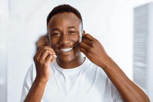 Toothcare And Hygiene. African American Guy Flossing Teeth Using Floss Smiling To Camera Standing In Modern Bathroom At Home. Healthy Teeth Cleaning Routine Concept photo
