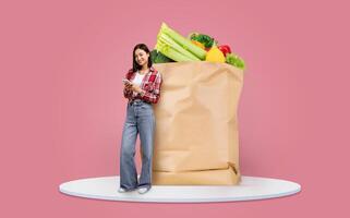 A customer stands beside a large paper bag filled with fresh vegetables and fruits while checking a list on her smartphone. Grocery shopping has become an important daily activity. photo